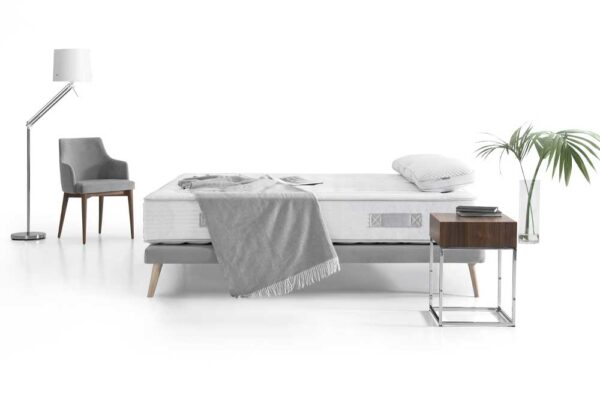 Configuración de dormitorio minimalista con una cama Colchón Karibian modelo Solid Firm cuidadosamente hecha, lámpara de pie, sillón, mesa auxiliar y planta en un espacio luminoso y limpio.