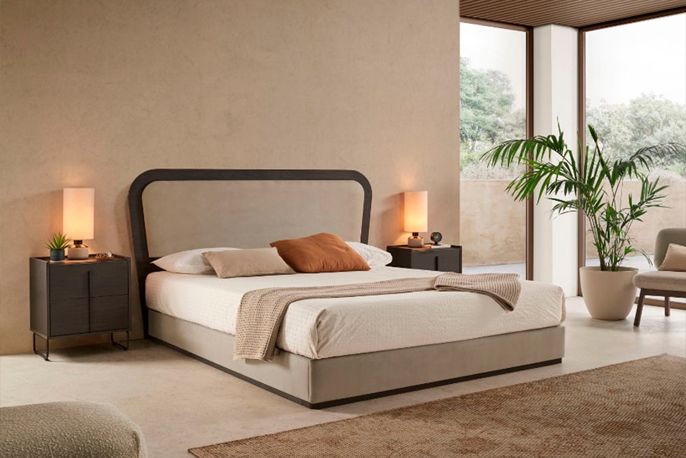Dormitorio moderno de diseño minimalista con cama grande, dos mesitas de noche con lámparas y zona de estar junto a la ventana. La pieza central es el suave Cabecero Astoria Soft, aportando un toque elegante.