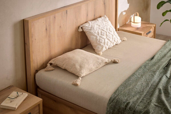 Una cama Cabecero modelo Kiara cuidadosamente hecha con almohadas y una manta texturizada en una habitación con iluminación tenue y muebles de madera.