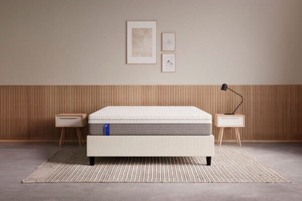 Un dormitorio minimalista con un Colchón EMMA Body Adapt Foam cuidadosamente elaborado con un marco simple, dos mesas de noche y una moderna lámpara de pie, todo ello contra una pared con paneles de madera en la parte inferior.