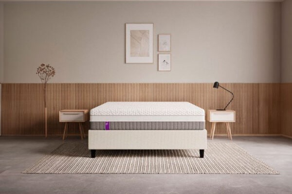 Un interior de dormitorio minimalista con una cama doble con colchón Colchón EMMA Fusion Fresh Hybrid, cabecera de madera, mesitas auxiliares y una alfombra beige.