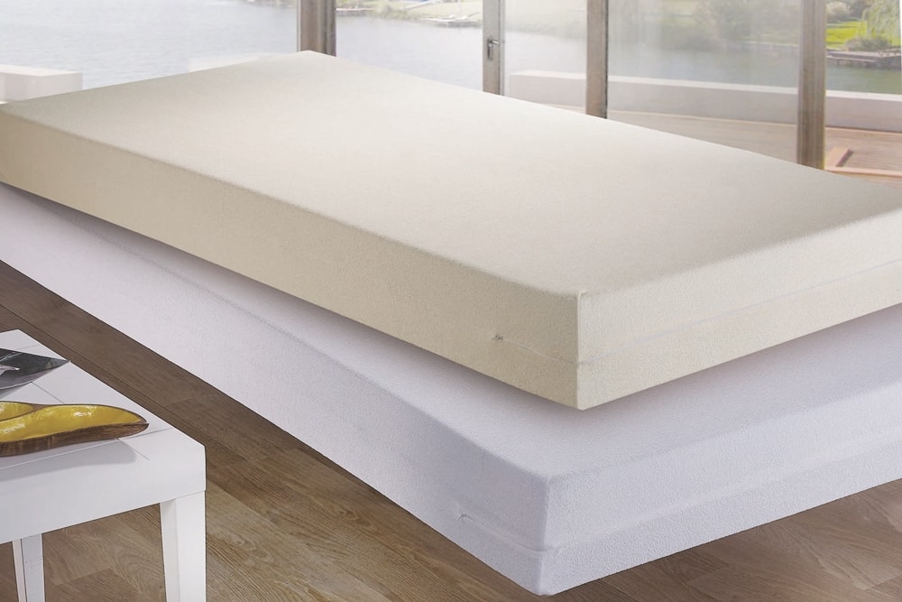 Funda de colchón Rizo de viscoelástica colocada sobre un colchón con una sábana blanca ajustada, con una vista escénica por la ventana en el fondo.