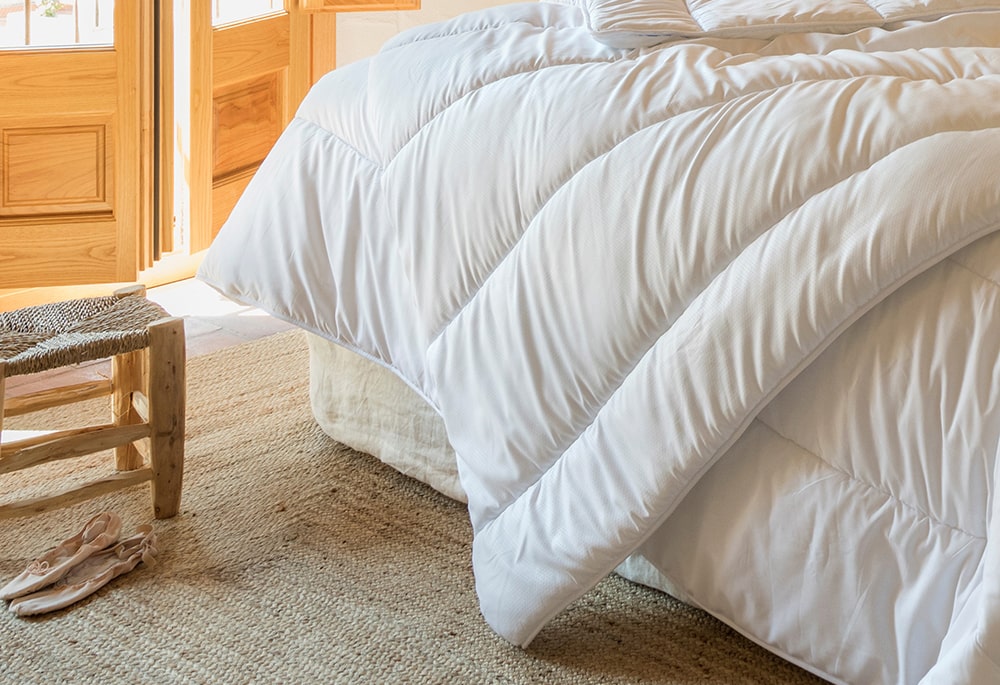 Una cama cuidadosamente hecha con plumón nórdico de fibra de aloe vera blanco en una habitación bien iluminada con detalles en madera y un par de sandalias en el suelo.