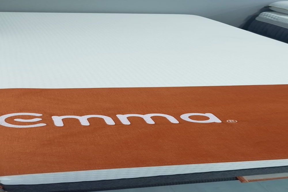 Imagen de un colchón con una funda naranja con la etiqueta "Emma" en el extremo de los pies. El colchón, identificado como Colchón EMMA Hybrid (copia), se encuentra colocado en un ambiente de sala de exposición.