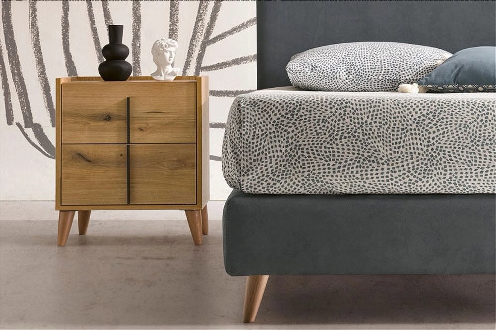 Un dormitorio moderno amueblado por Tori presenta una Mesita de noche de madera de Tori adornada con elementos decorativos, colocada junto a una cama con ropa de cama estampada y una cabecera tapizada en gris.