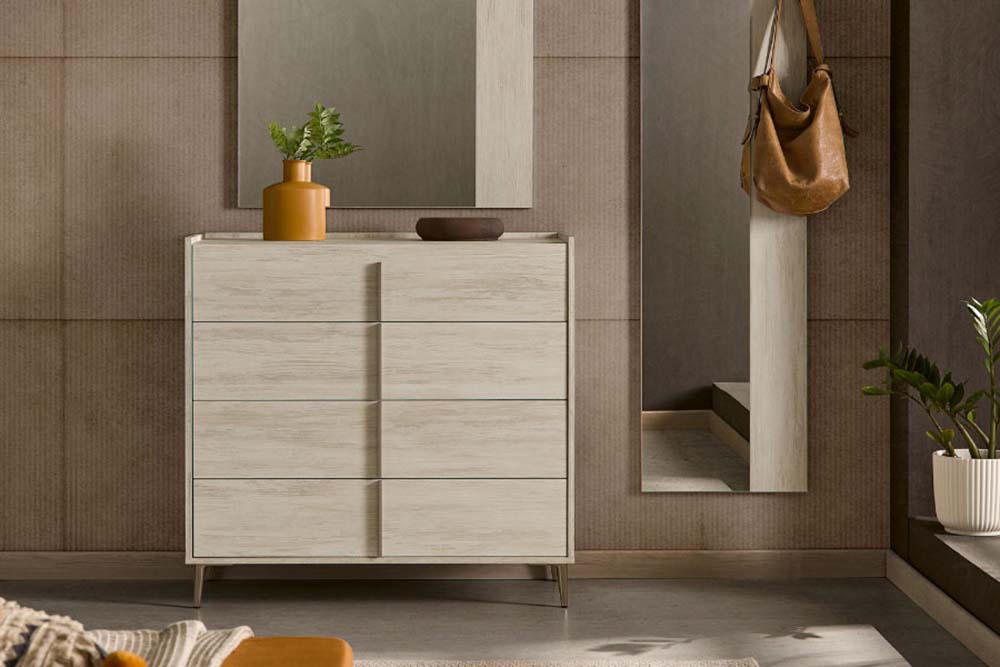 Una habitación minimalista cuenta con una cómoda de madera clara con seis cajones, una maceta, un jarrón marrón y un espejo al fondo junto a una bolsa marrón colgante.