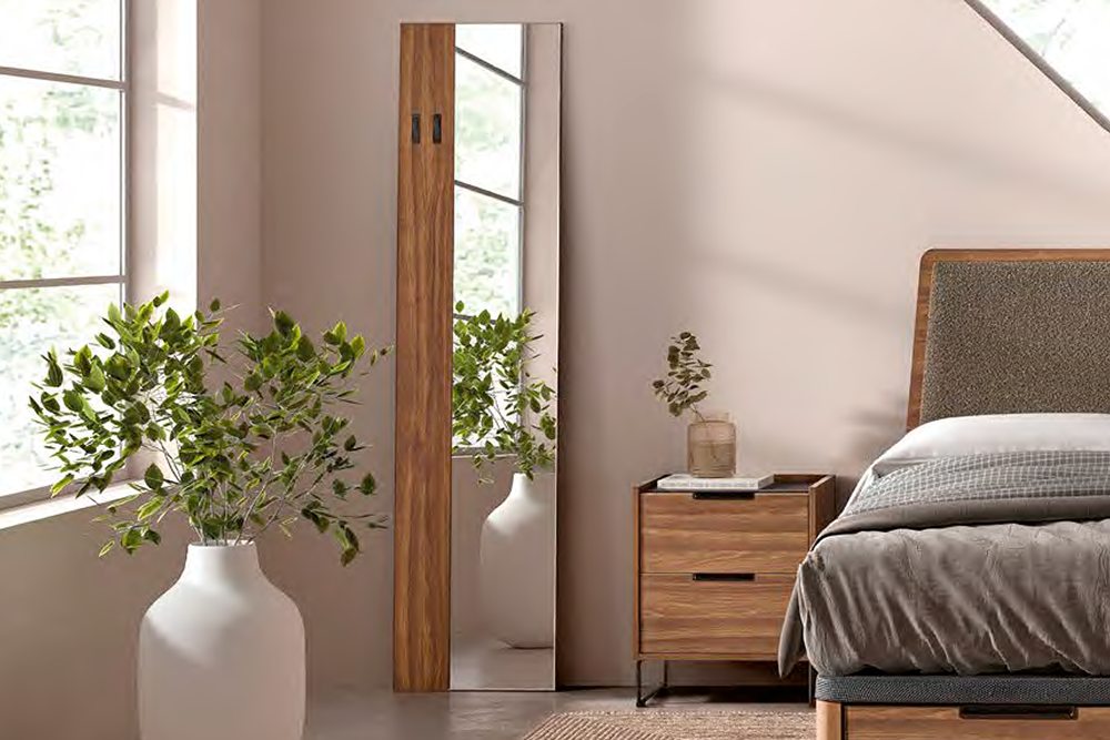 Un dormitorio con una mesilla de noche de madera, una cama con ropa de cama gris, un gran jarrón blanco con ramas verdes y el Espejo de pie Kiara reflejando parte de la habitación.