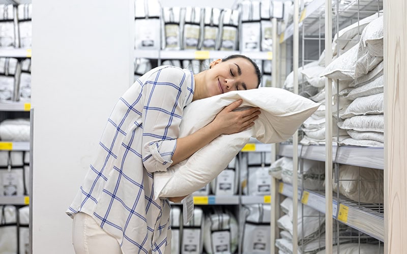 Una persona con una camisa a cuadros abraza amorosamente una almohada en el pasillo de una tienda, rodeada de estantes repletos de productos de cama envasados.