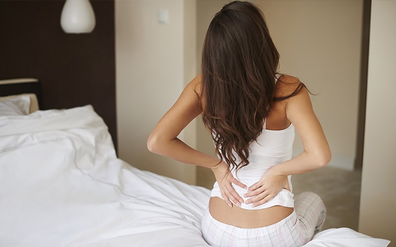 Una mujer está sentada en una cama, sujetándose la parte baja de la espalda con molestias, tal vez a causa del colchón. Lleva una camiseta blanca sin mangas y pantalones a cuadros.