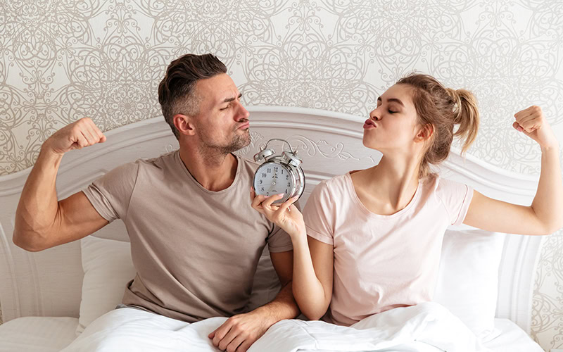 Un hombre y una mujer sentados en una cama flexionan sus brazos juguetonamente, mostrando los beneficios de un buen colchón con un reloj despertador sostenido entre ellos.