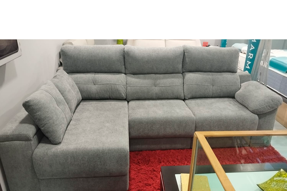 El Sofá Robinson exposición, en forma de L y color gris, se ubica sobre una alfombra roja junto a una mesa de madera con tapa de vidrio, creando un elegante ambiente de exposición.