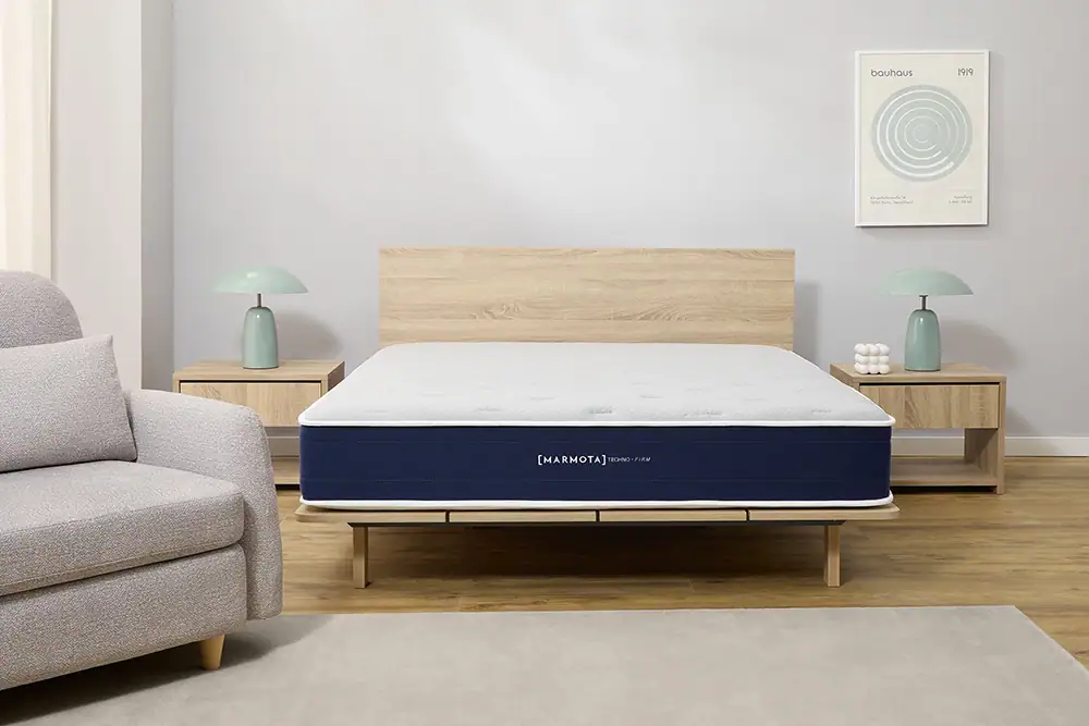 Una cama bien hecha con un colchón Colchón Marmota Techno Firm azul y blanco está centrada entre dos mesillas de noche de madera, cada una rematada con una lámpara verde, en un moderno dormitorio minimalista.